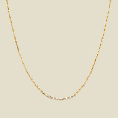 A delicate gold chain necklace featuring a short central crescent pattern with small clear baguette and round cut stones, displayed against a solid light beige background.