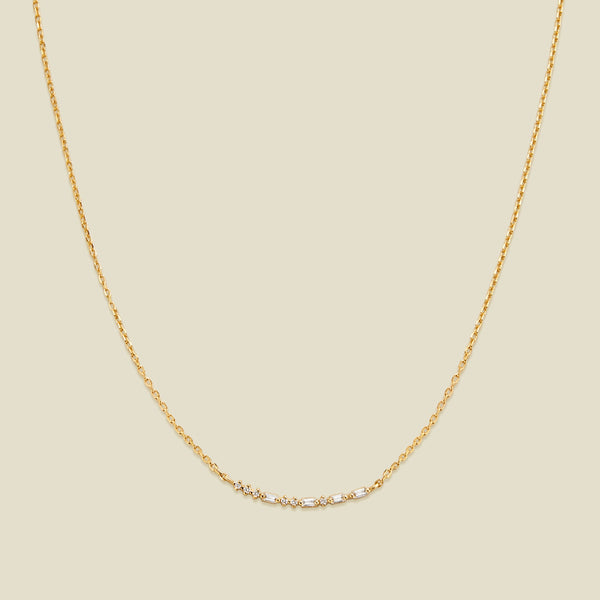 A delicate gold chain necklace featuring a short central crescent pattern with small clear baguette and round cut stones, displayed against a solid light beige background.