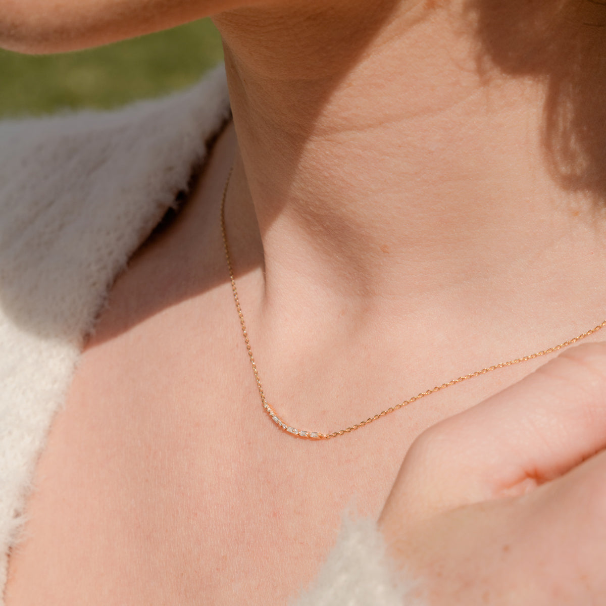 A close-up of a person's neck wearing a delicate gold chain necklace featuring a short central crescent pattern with small clear baguette and round cut stones.