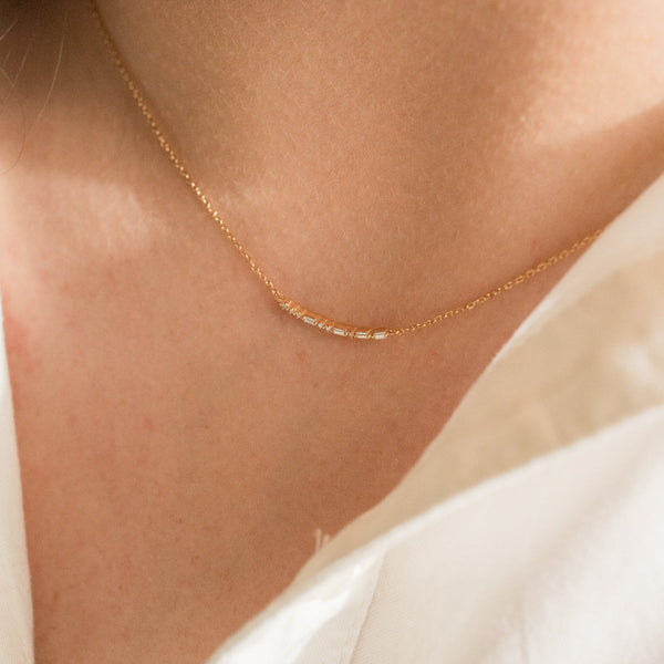 A close-up of a person's neck wearing a delicate gold chain necklace featuring a short central crescent pattern with small clear baguette and round cut stones.