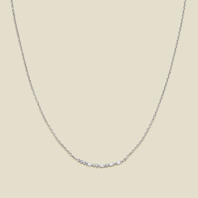 A delicate silver chain necklace featuring a short central crescent pattern with small clear baguette and round cut stones, displayed against a solid light beige background.