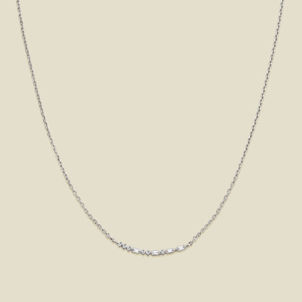 A delicate silver chain necklace featuring a short central crescent pattern with small clear baguette and round cut stones, displayed against a solid light beige background.