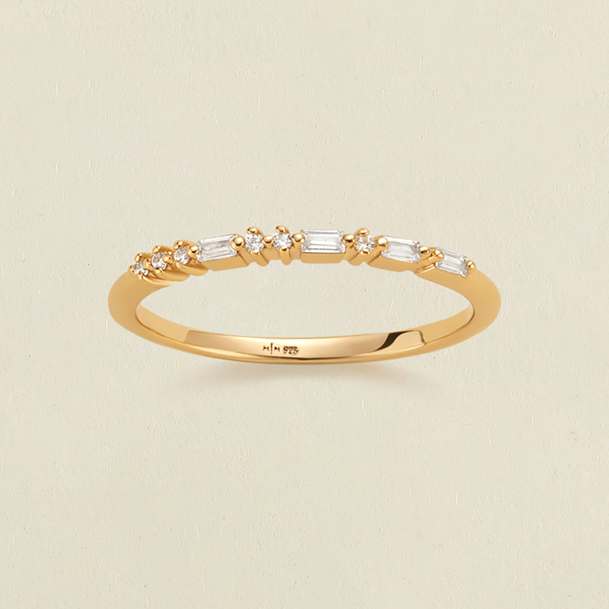 A delicate yellow gold ring with a row of clear baguette and round-cut stones in a morse code pattern, photographed on a light beige textured background.