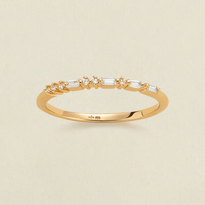 A delicate yellow gold ring with a row of clear baguette and round-cut stones in a morse code pattern, photographed on a light beige textured background.