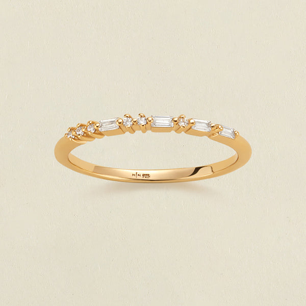 A delicate yellow gold ring with a row of clear baguette and round-cut stones in a morse code pattern, photographed on a light beige textured background.