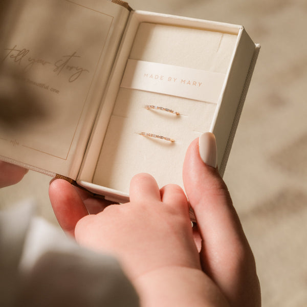 A person holding a cream colored jewelry display box with a necklace wrap reading 'MADE BY MARY'. Inside are two delicate gold rings, each with a different pattern of baguette and round cut clear gemstones.