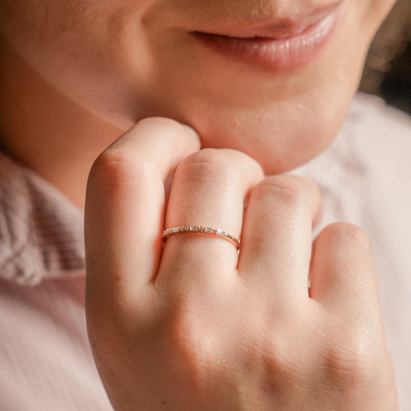 A person resting their hand on their chin showcasing a delicate gold ring. The ring has a single row of clear baguette and round cut gemstones in a morse code pattern.