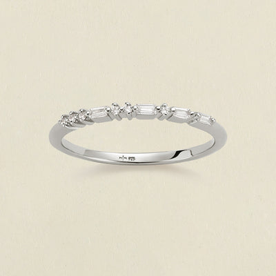 A delicate silver ring with a row of clear baguette and round-cut stones in a morse code pattern, photographed on a light beige textured background.