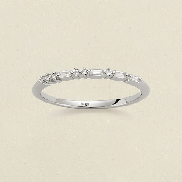 A delicate silver ring with a row of clear baguette and round-cut stones in a morse code pattern, photographed on a light beige textured background.