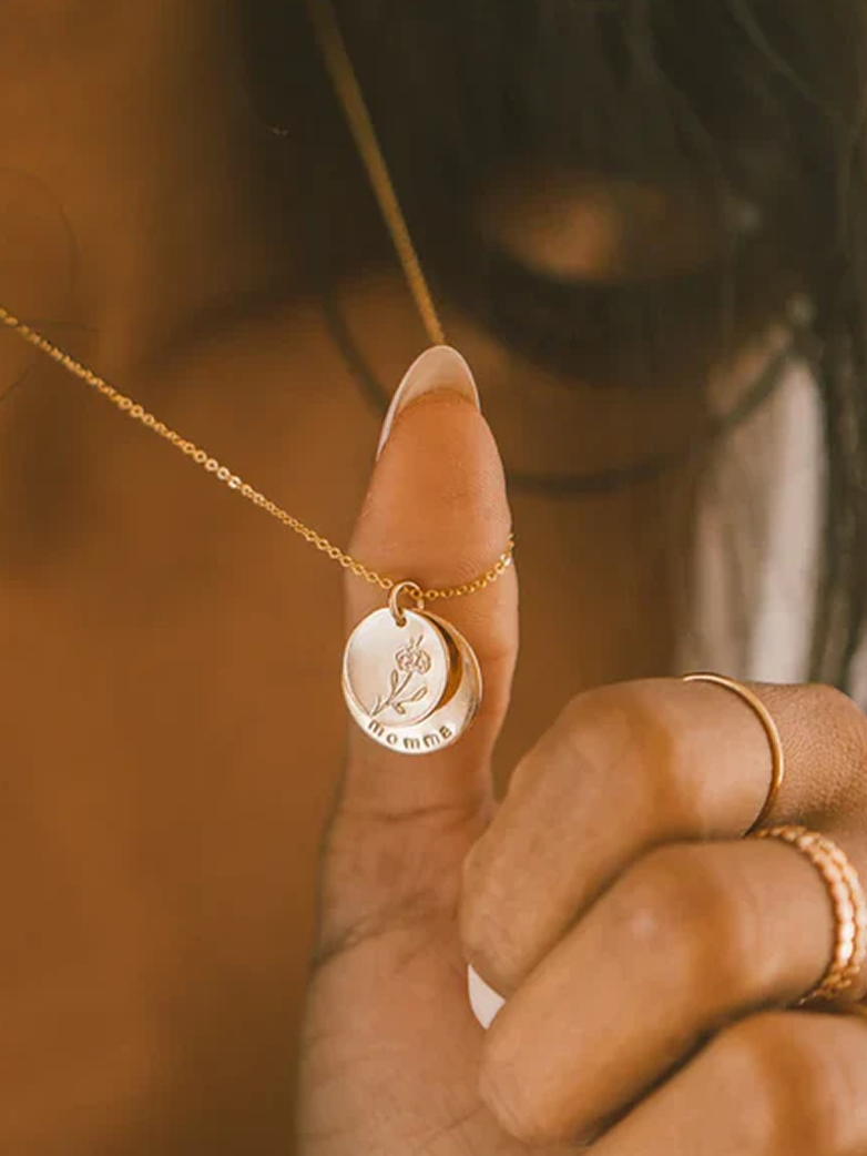 A close up lifestyle shot of a person's neck wearing a dainty gold chain necklace with a layered, round charm with two discs. The top disc features a floral design, and the larger bottom disc has the word "momma".