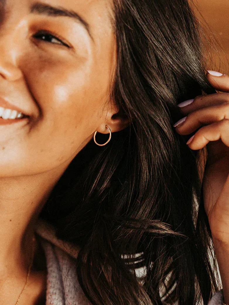 Close-up of a smiling woman wearing a thin gold hoop earring, and holding back her long dark hair with her hand.