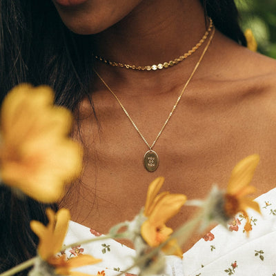 A woman wearing a gold choker with round flat gold disc links and a longer delicate gold pendant chain with an engraved oval disc charm. There are yellow flowers in the foreground.