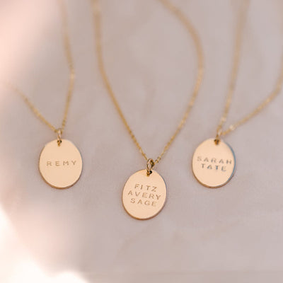 Three delicate gold pendant necklaces with flat oval discs. Each disc is engraved with either 'REMY', 'FITZ, AVERY, SAGE', or 'SARAH, TATE'.  