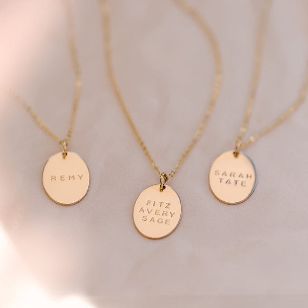 Three delicate gold pendant necklaces with flat oval discs. Each disc is engraved with either 'REMY', 'FITZ, AVERY, SAGE', or 'SARAH, TATE'.  