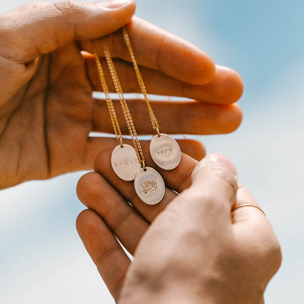 A person's hands holding three gold delicate pendant chain necklaces with flat oval charms, each with a different engraving. Two have text and one has a floral motif.