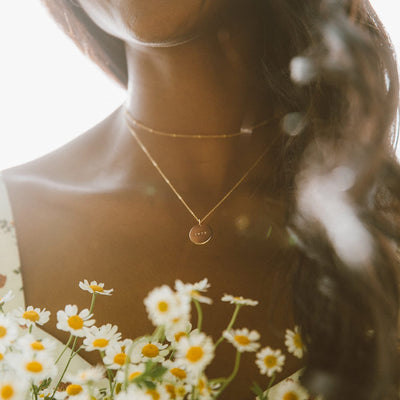 A person holding small white daisies, wearing a dainty gold choker necklace and a delicate pendant chain with a flat oval disc.