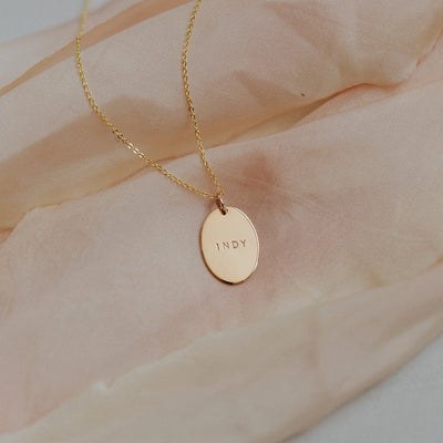 A delicate gold pendant chain necklace with a flat oval disc charm, engraved with the name 'INDY'. The necklace is laying on a blush colored fabric.