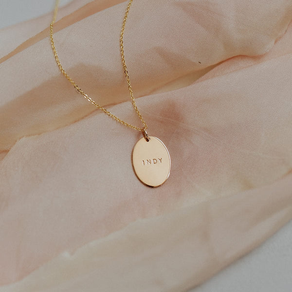 A delicate gold pendant chain necklace with a flat oval disc charm, engraved with the name 'INDY'. The necklace is laying on a blush colored fabric.