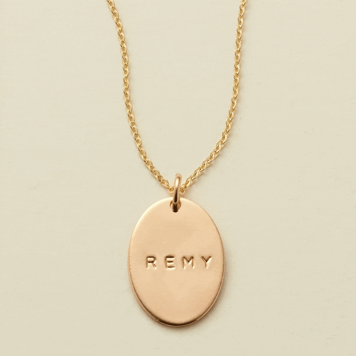 A gif of a delicate gold chain pendant necklace with a flat gold oval charm. The charm is engraved, and the engraving changes between different names.