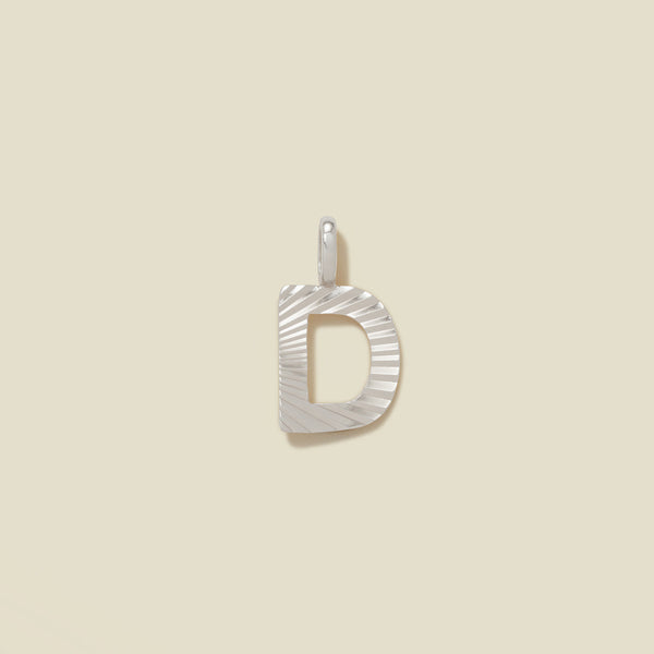 A small silver D-shaped charm with a textured, sunburst striped pattern, shown on a solid light beige background.