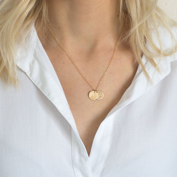 A close-up of a woman in a white button-up top wearing a delicate gold pendant chain necklace with two round flat disc charms, both engraved with the letter 'A'.