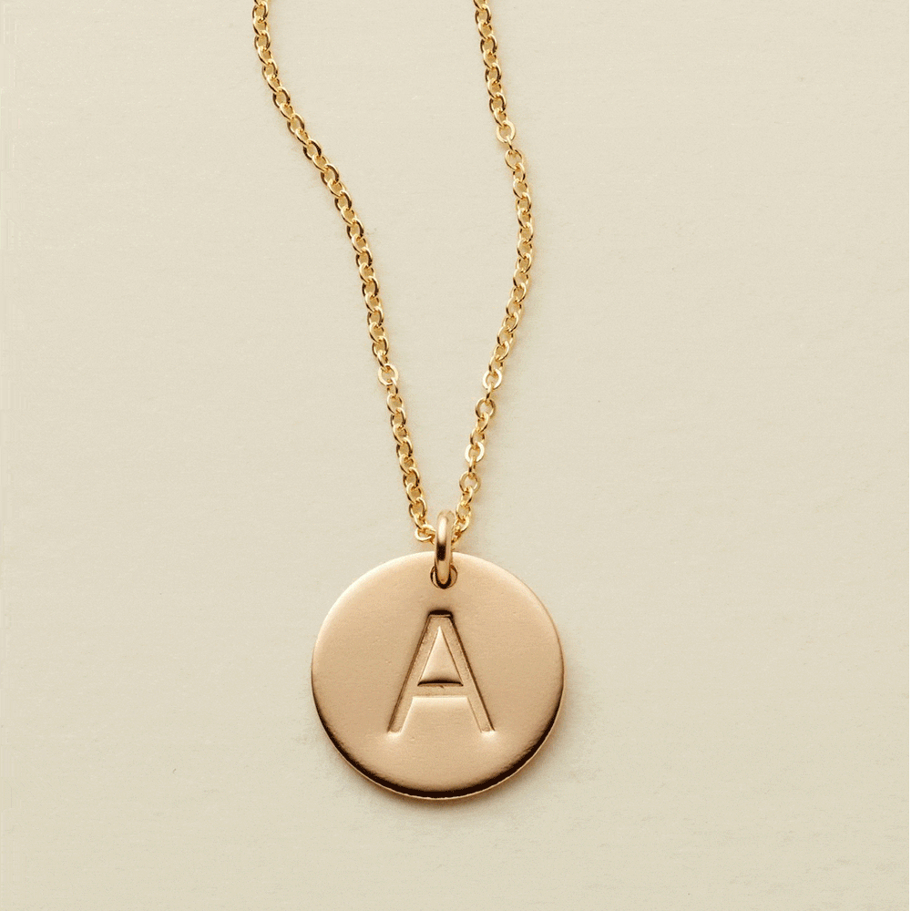 A gif of a delicate gold chain necklace with a round flat disc charm engraved with the letter 'A'. One by one more engraved discs with letters get added to the chain, up to three.