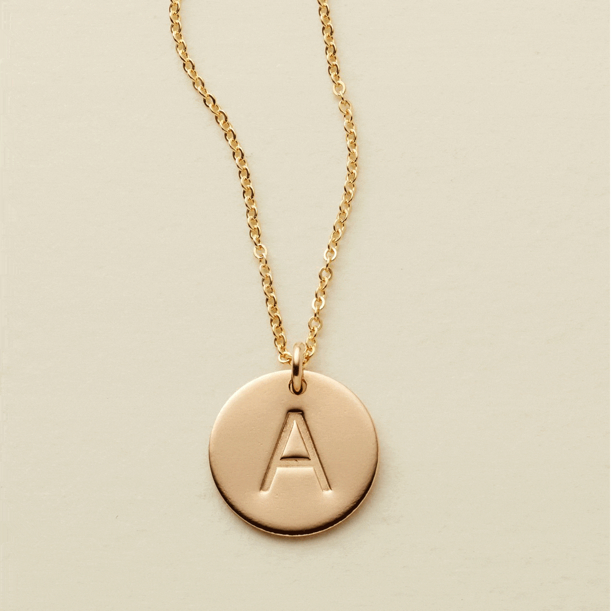 A gif of a delicate gold chain necklace with a round flat disc charm engraved with the letter 'A'. One by one more engraved discs with letters get added to the chain, up to three.