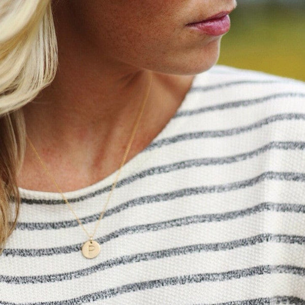 A woman in a white and navy blue striped top wearing a delicate gold pendant chain necklace with a round disc charm engraved with the letter 'P'.