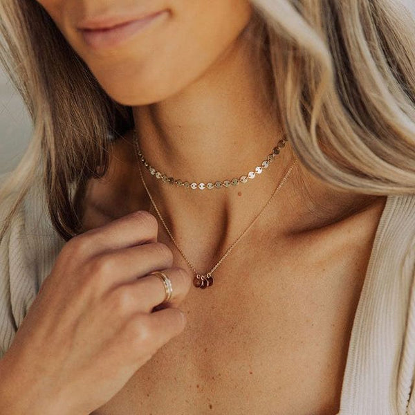 A close-up of a woman's neck wearing a gold choker necklace made up of flat round discs and a longer delicate gold pendant chain with three small round disc charms.