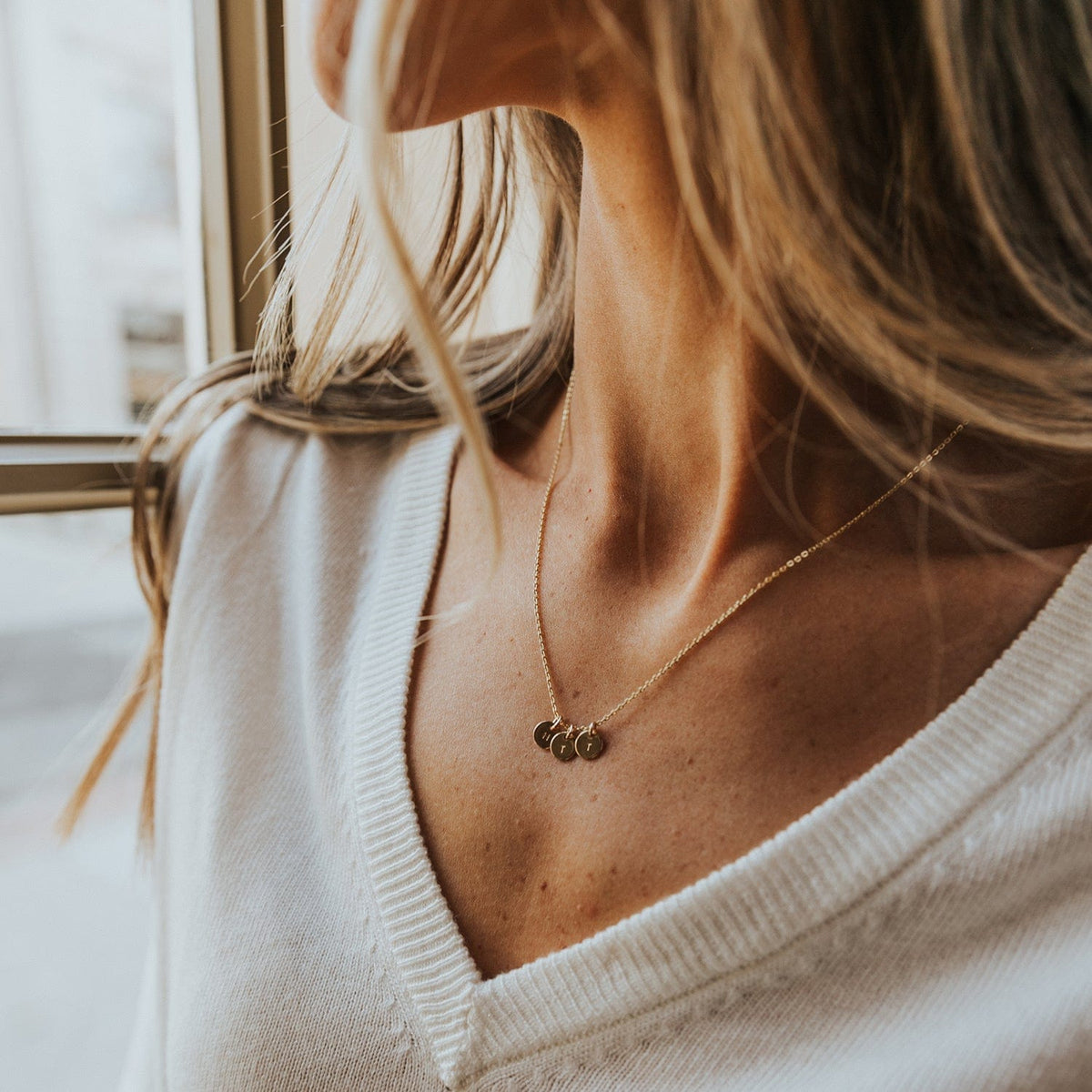 A woman in a white knit v-neck sweater wearing a delicate gold pendant necklace with three round disc charms, each engraved with an initial.