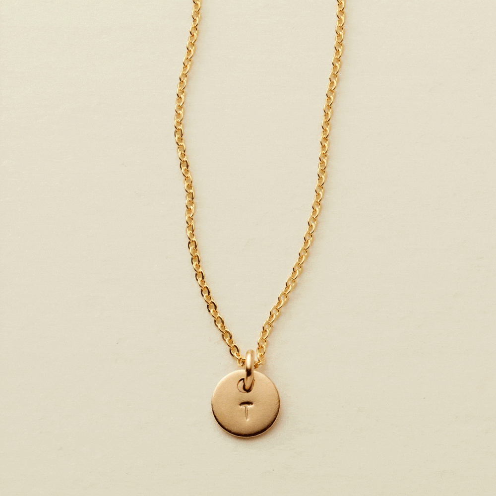 A gif of a delicate gold pendant chain necklace with a small round flat disc charm engraved with the letter 'T'. One by one more discs are added, each with an engraved letter up to five discs.