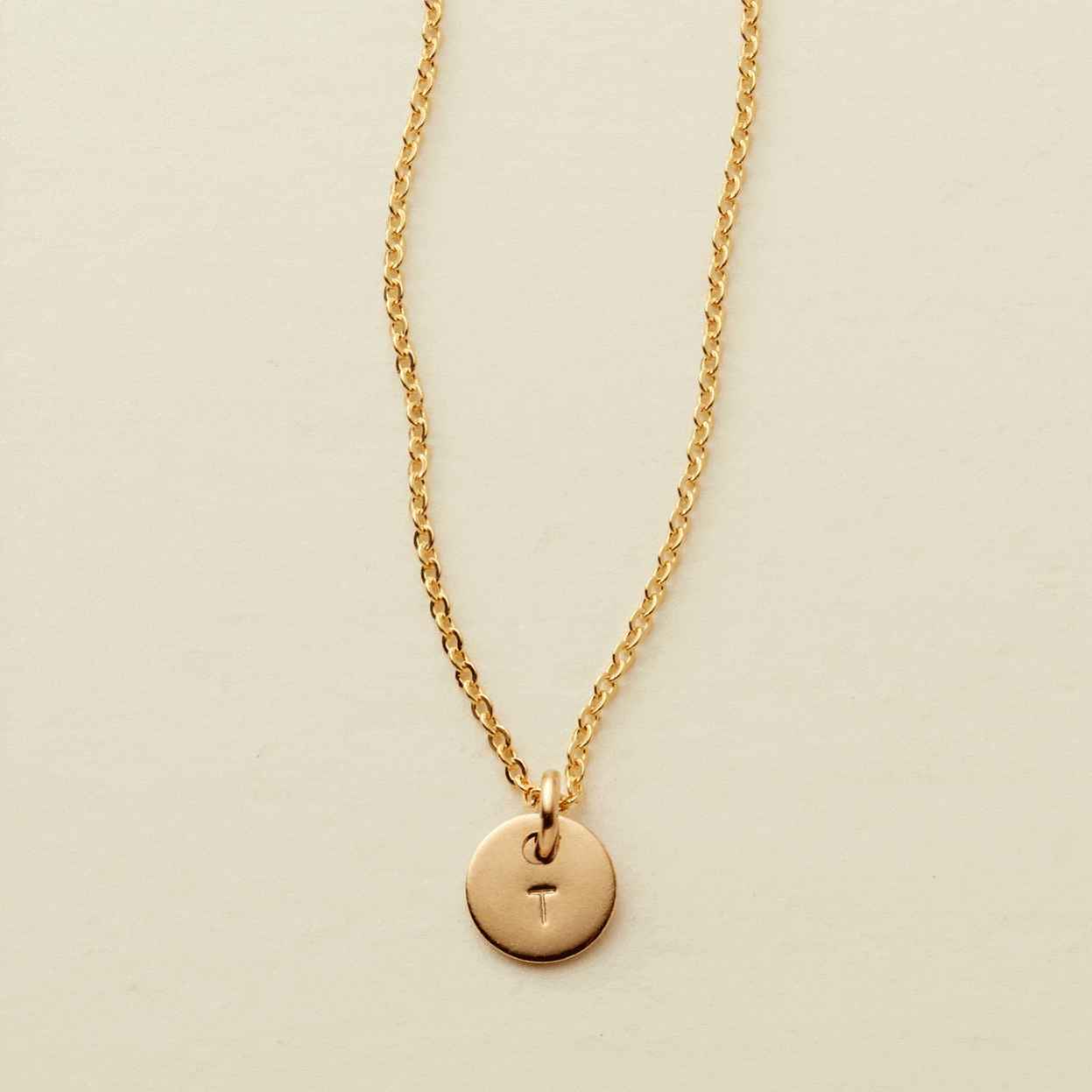 A gif of a delicate gold pendant chain necklace with a small round flat disc charm engraved with the letter 'T'. One by one more discs are added, each with an engraved letter up to five discs.