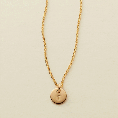 A gif of a delicate gold pendant chain necklace with a small round flat disc charm engraved with the letter 'T'. One by one more discs are added, each with an engraved letter up to five discs.