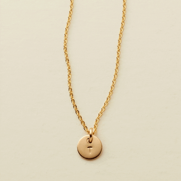 A gif of a delicate gold pendant chain necklace with a small round flat disc charm engraved with the letter 'T'. One by one more discs are added, each with an engraved letter up to five discs.