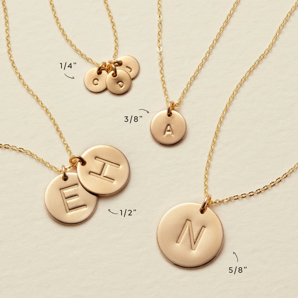 Four gold pendant chain necklaces laying on a textured light beige surface, each with a different size of round disc charms engraved with letters. They are all labeled with sizes: 1/4", 3/8", 1/2", and 5/8".