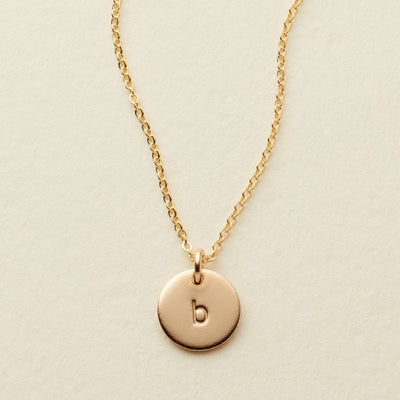 A delicate gold pendant chain necklace with a small round flat disc charm engraved with the letter 'b', shown on a textured light beige surface.