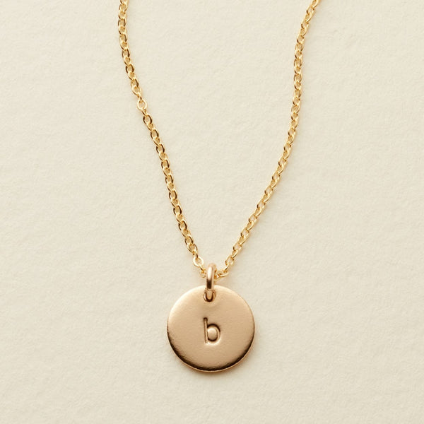 A delicate gold pendant chain necklace with a small round flat disc charm engraved with the letter 'b', shown on a textured light beige surface.