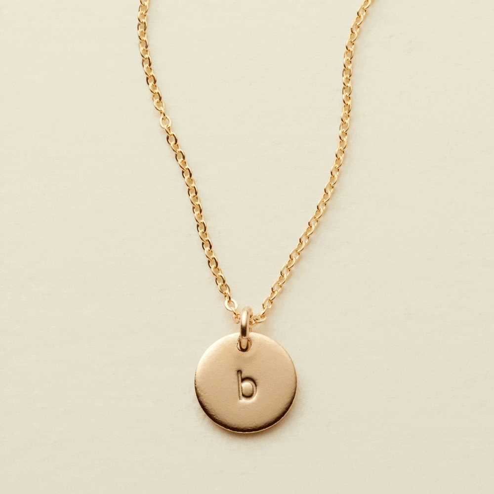 A gif of a delicate gold pendant chain necklace with a round flat disc charm engraved with the letter 'b'. One by one additional engraved discs get added to the necklace, up to four.