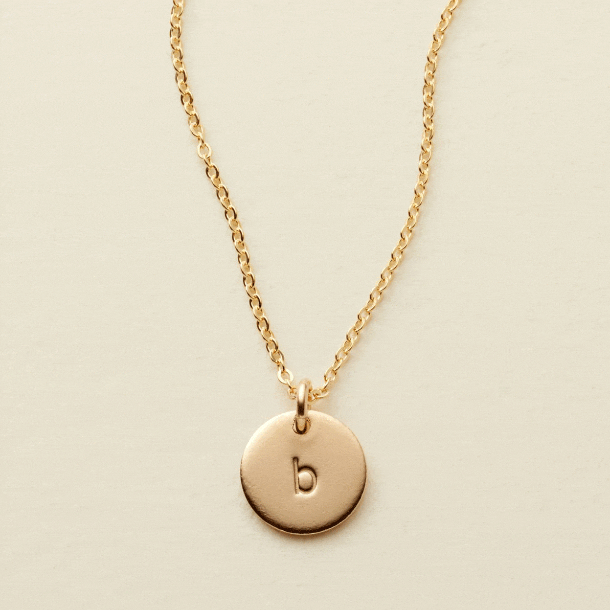 A gif of a delicate gold pendant chain necklace with a round flat disc charm engraved with the letter 'b'. One by one additional engraved discs get added to the necklace, up to four.