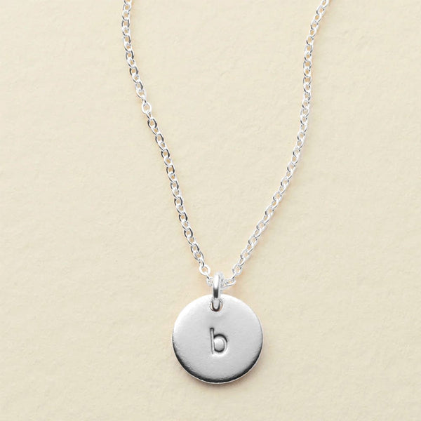 A delicate silver pendant chain necklace with a flat round disc charm engraved with the letter 'b', shown on a textured light beige surface.