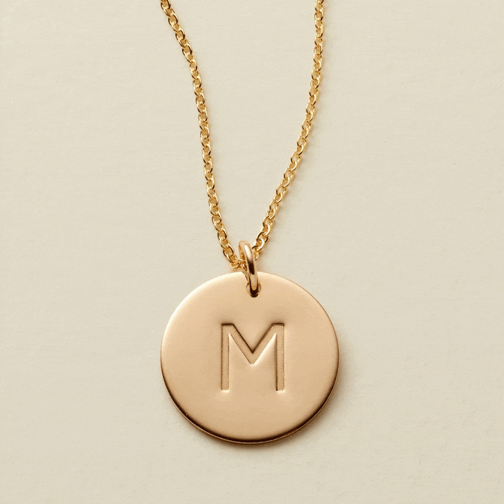 A gif of a delicate gold pendant necklace with a round flat disc engraved with the letter 'M'. It then switches to two round discs engraved with the letters 'N' and 'S'.