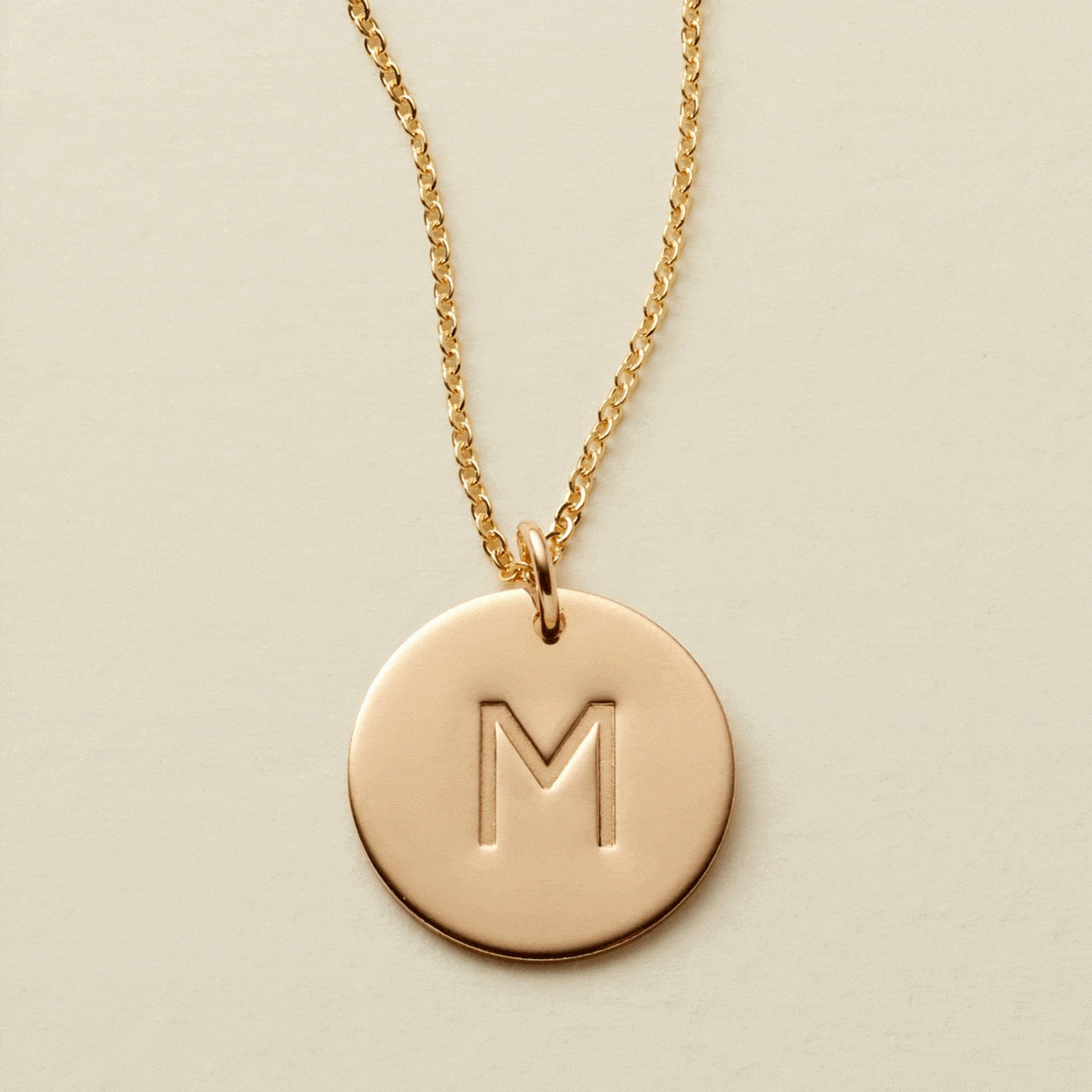 A gif of a delicate gold pendant necklace with a round flat disc engraved with the letter 'M'. It then switches to two round discs engraved with the letters 'N' and 'S'.