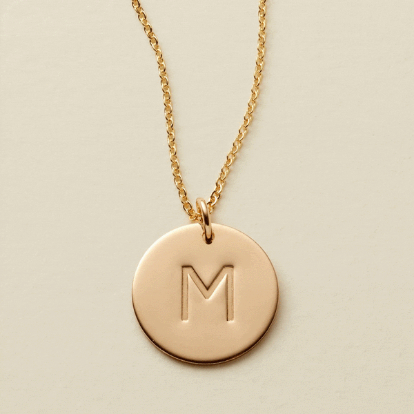 A gif of a delicate gold pendant necklace with a round flat disc engraved with the letter 'M'. It then switches to two round discs engraved with the letters 'N' and 'S'.