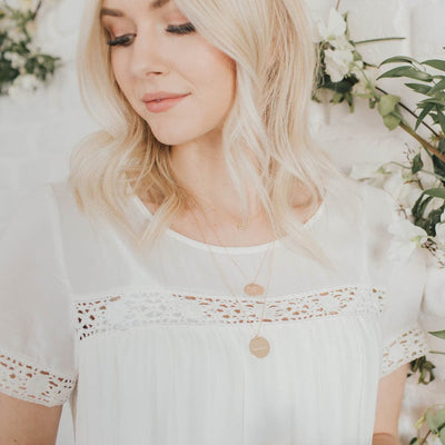 A blonde woman in a dainty white top with green and white foliage behind her, wearing two long delicate pendant necklaces with round flat disc charms.