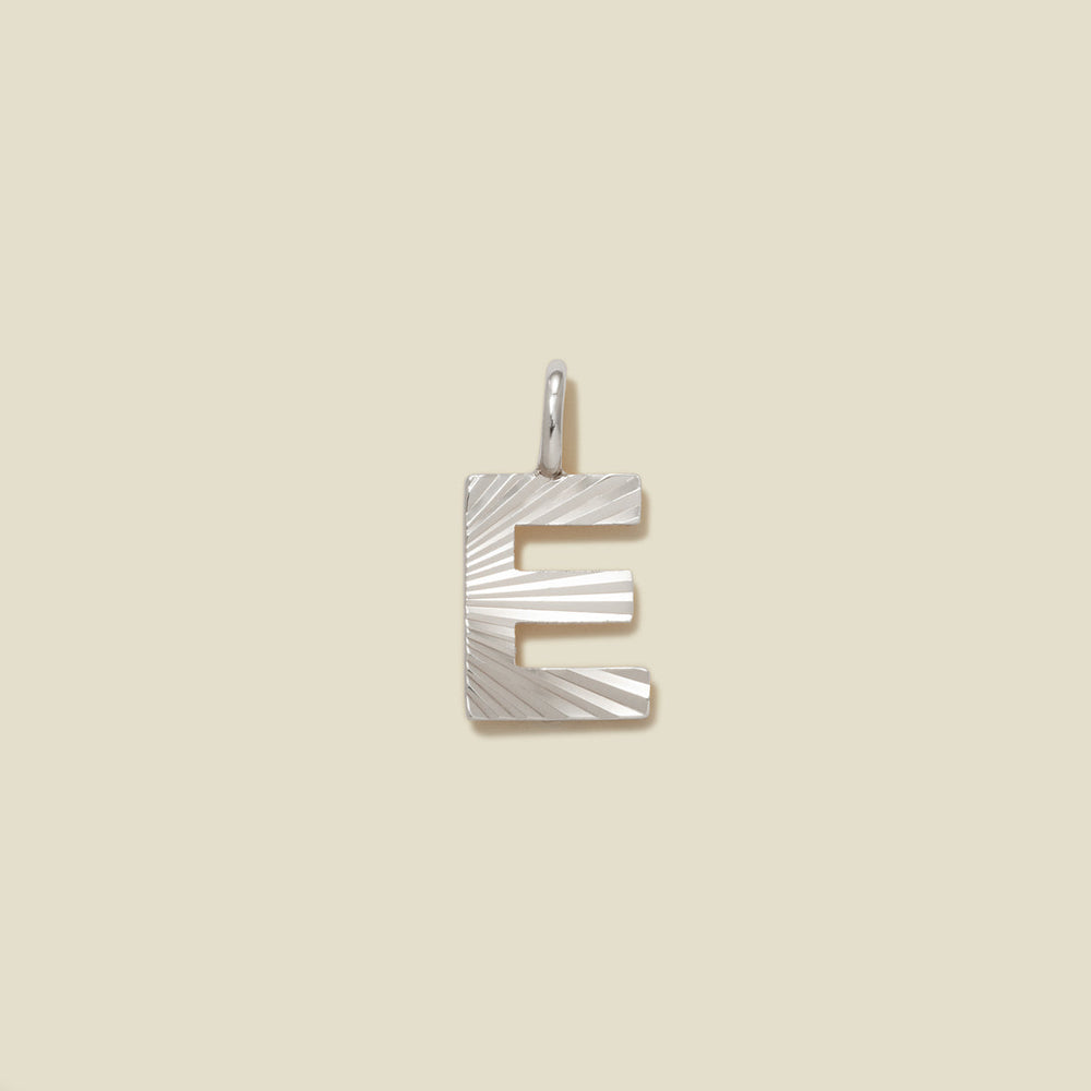 A small silver E-shaped charm with a textured, sunburst striped pattern, shown on a solid light beige background.