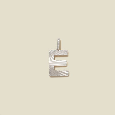 A small silver E-shaped charm with a textured, sunburst striped pattern, shown on a solid light beige background.