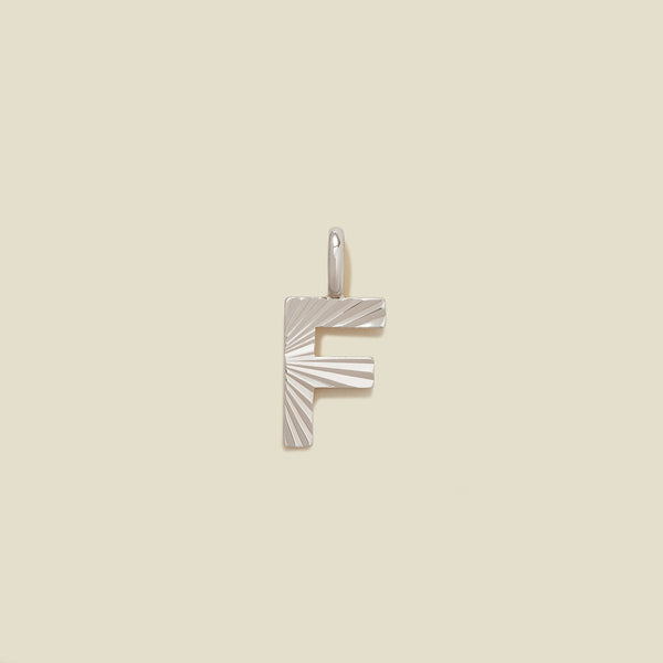 A small silver F-shaped charm with a textured, sunburst striped pattern, shown on a solid light beige background.