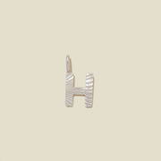 A small silver H-shaped charm with a textured, sunburst striped pattern, shown on a solid light beige background.