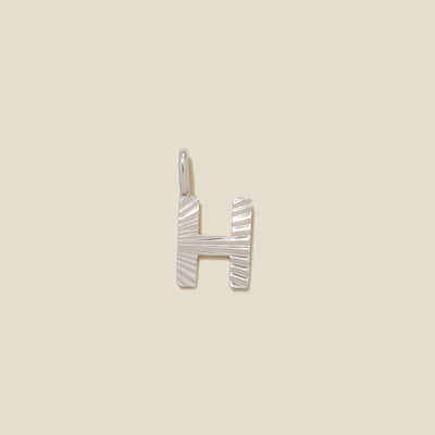 A small silver H-shaped charm with a textured, sunburst striped pattern, shown on a solid light beige background.