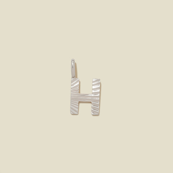 A small silver H-shaped charm with a textured, sunburst striped pattern, shown on a solid light beige background.